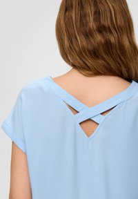 Vrouw met golvend bruin haar die een lichtblauw topje draagt met een gekruiste schouderband en een V-vormige uitsnijding op de bovenrug.
