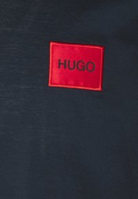 Tissu bleu marine foncé avec un petit patch carré rouge portant le texte noir "HUGO" brodé au centre.