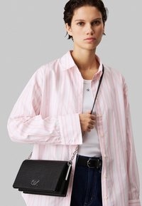 Svart lærveske med teksturert overflate, sølvfargede detaljer og kjedehåndtak, holdt av en person iført en rosa stripete skjorte og jeans.