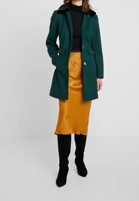 Cappotto di lana verde con collo in pelliccia nera, tasche frontali e bottoni dorati, indossato sopra una gonna in satin gialla elegante e stivali neri fino al ginocchio.