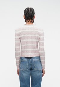 Personu ar pinumiem matiem, ģērbtu rozā un baltā svītrainā adīta džempera un brīvu, zilu džinsu, redz no mugurpuses.