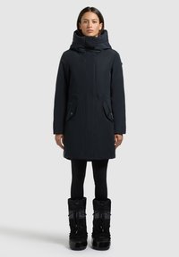 Sort parka med høj krave og lynlås, der har to forlommer og en tætsiddende silhuet, parret med knæhøje sorte støvler.