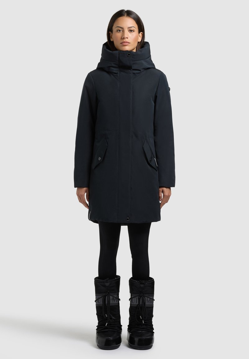 Sort parka med høj krave og lynlås, der har to forlommer og en tætsiddende silhuet, parret med knæhøje sorte støvler.