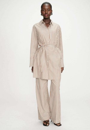 Femme debout portant une tunique boutonnée à rayures verticales beige et blanc avec un pantalon palazzo assorti et des talons ouverts noirs.