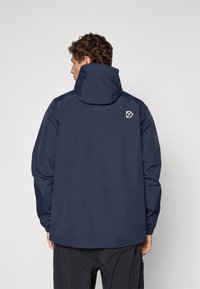 Homme aux cheveux bouclés portant une veste à capuche navy avec un petit logo géométrique blanc dans le haut du dos, sur fond uni.
