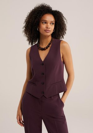 Kvinde med krøllet hår iført en mørkelilla vest med knapper og matchende bukser, accessoriseret med en stor mørk perlehalssmykke og creoløreringe.