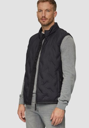 Schwarze, gesteppte Pufferweste mit hohem Kragen und Reißverschluss vorne. Verfügt über einen tonalen Logostreifen entlang des Reißverschlusses. Trägt man über einem grauen Langarmshirt.