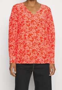 Rote und rosa Langarmbluse mit V-Ausschnitt, abstraktem Muster, strukturiertem Stoff und lässiger Passform. Kombiniert mit schwarzen Hose.