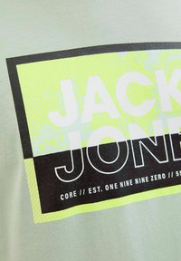 Lys grøn t-shirt med sort og neongul grafisk tekst med striber og et rektangulært design. Teksten inkluderer "JACK" og "JONES."