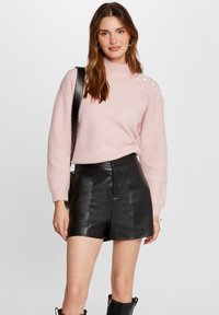 Różowy sweter w prążki z wysokim kołnierzem, z guziczkami na ramionach, zestawiony z czarnymi skórzanymi szortami i kozakami sięgającymi do kolan.