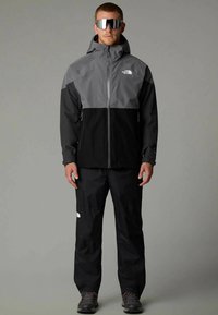 Veste imperméable en gris et noir, avec capuche, fermeture éclair sur le devant et logo. Associée à un pantalon assorti et des chaussures robustes.