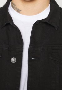 Svart denimjacka med framfickor, silverfärgad knapprad och stickade detaljer. Buren över en vit T-shirt som visar en enkel textur.