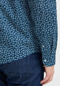 Camicia blu navy con bottoni, caratterizzata da un motivo di foglie bianche, con orlo dritto, maniche lunghe e sfondo in denim blu.