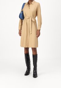 Beżowa sukienka zapinana na guziki z paskiem w talii, z długimi rękawami i gładką teksturą, w połączeniu z wysokimi na kolana czarnymi skórzanymi botkami.