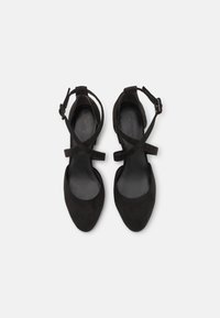 Paire de chaussures pour femmes en daim noir avec des bouts pointus et des brides croisées à la cheville avec fermeture par boucle sur fond blanc.