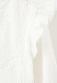 Bílá halenka s texturovanou látkou s malými vystouplými tečkami. Má krajkový lem a volánové detaily na rukávech, které dodávají jemný design.