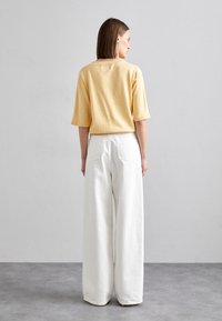 Krátký rukáv žlutého pleteného topu spojený s volnými bílými kalhotami, obě mají hladké textury a minimalistický design.