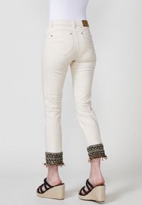 Pantalones color crema con puños bordados y con cuentas. Presenta un diseño ajustado, bolsillos traseros y una etiqueta de logo de cuero en la cintura.