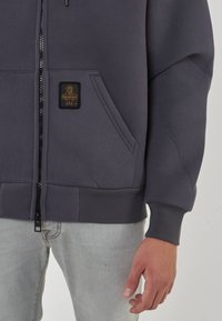 Felpa grigia con zip, dotata di una grande tasca frontale, polsini e orlo a costine. Presenta una piccola toppa con logo giallo e nero sulla tasca.