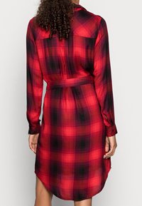 Czerwona i czarna koszulowa sukienka w kratę z długimi rękawami, paskiem w talii i zaokrąglonym dołem. Wykonana z miękkiego, lekkiego materiału. Widoczne guzikowe mankiety.
