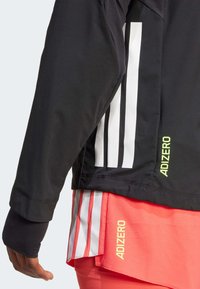 Chaqueta deportiva negra con acentos en rayas blancas y la marca "ADIZERO" en verde neón. Cuenta con un alto cuello y tejido texturizado.