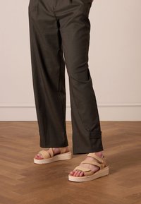 Beige platform sandals with cross-strap design and textured straps, worn with dark gray, loose-fitting trousers. Smooth wooden floor backdrop.