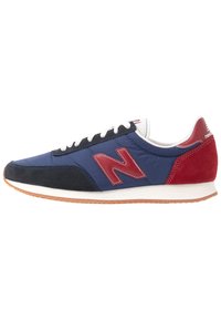 Chaussure de sport bleue et rouge avec une tige en daim et nylon, des lacets blancs et une semelle en caoutchouc gomme. Présente un logo "N" rouge proéminent.