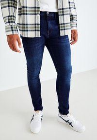 Mörkblå slim-fit jeans med en slät textur, i kombination med vita sneakers med marinblå detaljer. Skjortan har ett rutig mönster i vitt och grönt.