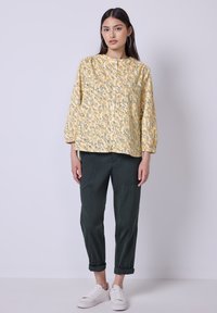 Blouse florale jaune avec un col rond et des manches trois-quarts, associée à un pantalon tapered vert foncé et des baskets blanches.