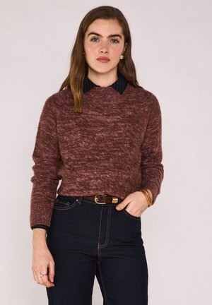 Femme aux longs cheveux bruns portant un pull bordeaux sur une chemise noire à col, un jean foncé, une ceinture marron et des bracelets, debout avec une main dans la poche.