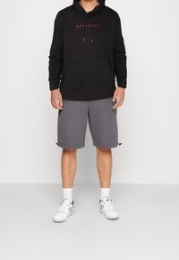 Persona con sudadera negra con bolsillo frontal, pantalones cortos grises hasta la rodilla con dobladillos ajustables, calcetines blancos y zapatillas blancas de pie.