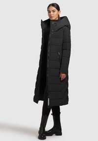Hosszú fekete puffernadrág kapucnival, steppelt textúrával, elöljáró cipzárral, oldalsó zsebekkel és logós címkével az alján.
