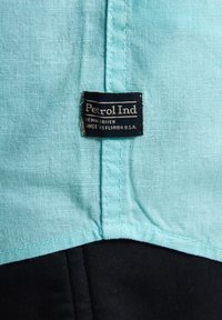 Een stoffen label van Petrol Ind, bevestigd aan lichtblauwe stof met een gladde textuur, boven een zwart gedeelte, met een bedrijfslogo.