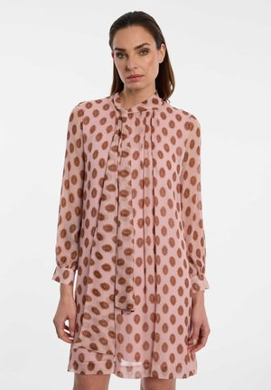 Vrouw draagt een lange mouwen roze jurk met bruine cirkelvormige patronen en een strikdetail bij de kraag, staand tegen een effen witte achtergrond.