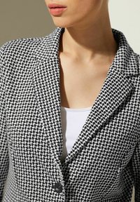 Blazer à motifs noir et blanc avec un revers cranté et une fermeture à bouton unique, porté sur un débardeur blanc ; tissu texturé.