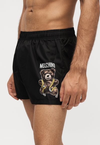 Άνδρας με μαύρα σορτς με κείμενο Moschino και γραφικό αρκουδάκι τυλιγμένο σε κίτρινη μεζούρα στην αριστερή πλευρά.