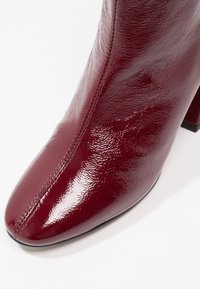 Bottine en cuir verni rouge foncé brillant avec bout arrondi, coutures visibles et talon bloc sur fond blanc.