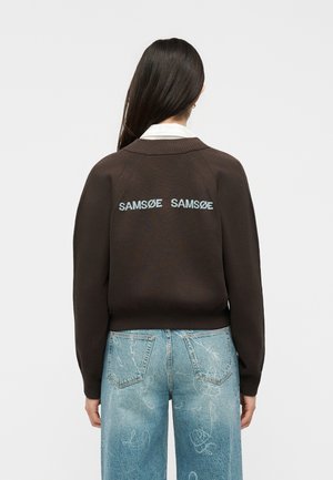 Persoon met lang donker haar die een bruine trui draagt met de tekst "SAMSØE SAMSØE" op de rug en lichtblauwe spijkerbroek met witte krabbels.