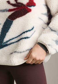 Fleecový pulóver v krémovej farbe s kvetinovým vzorom v odtieňoch bordovej a tyrkysovej. Mäkká textúra s bielym lemom na spodnom okraji.