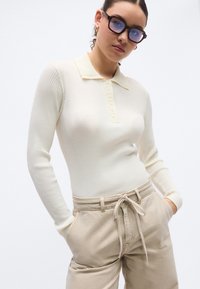 Μπλούζα πόλο με μακριά μανίκια σε κρεμ χρώμα με ριγέ υφή, γιακά με κυματιστό σχέδιο και κουμπιά στο μπροστινό μέρος, συνδυασμένη με μπεζ παντελόνια ψηλόμεσα με ζώνη στη μέση.