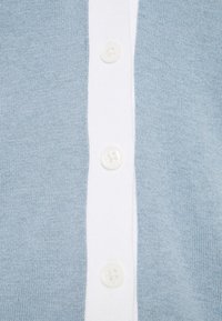 Ljusblått stickat tyg med en vertikal vit rand som pryds av tre jämnt fördelade vita knappar i mitten.