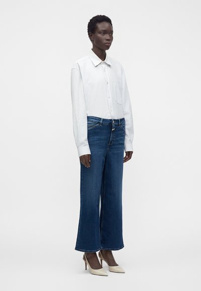 Femme portant une chemise blanche boutonnée, un jean bleu à jambes larges et des talons beiges, debout devant un fond clair uni.