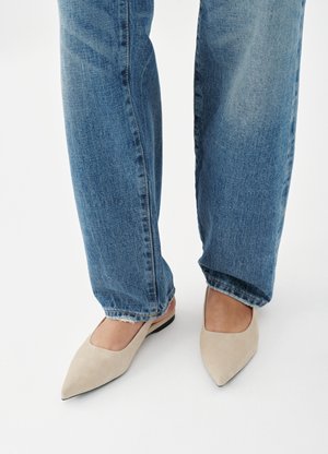 Person iført blå jeans og beige spidse flade sko, stående på et hvidt gulv.