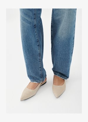 Žmogus dėvi mėlynas džinsines kelnes ir smėlio spalvos smailią butų batų porą, stovintis ant balto grindų paviršiaus.