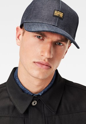Dunkelblaue Denim-Basecap mit strukturierter Krone und flachem Schirm; verfügt über ein goldenes Logopatch auf der Vorderseite. Schwarze Jacke und blauer Kragen sichtbar.