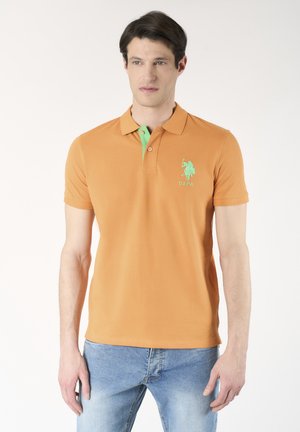 Uomo con polo arancione a maniche corte con logo verde e jeans blu, in piedi davanti a uno sfondo bianco uniforme.