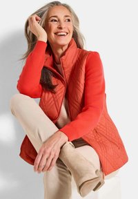 Lächelnde reife Frau mit grauen Haaren, die eine rote gesteppte Weste, ein rotes Langarmshirt, beige Hosen und beiger Wildlederschuhe trägt, entspannt sitzt.