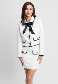 Biała tweedowa marynarka z czarnymi wykończeniami, z kokardą, złotymi guzikami oraz dwiema przednimi kieszeniami. W komplecie dopasowana spódnica.