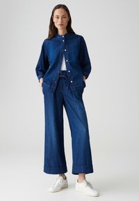 Dunkelblaue Jeansjacke und weite Hose. Die Jacke hat Knöpfe und einen Kragen, während die Hose einen Kordelzug und einen umgenähten Saum hat.