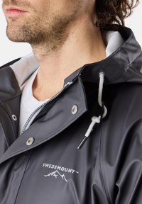 Homme portant une veste de pluie Swedemount gris foncé avec un cordon blanc et des boutons pression en argent, montrant le cou et le bas du visage.